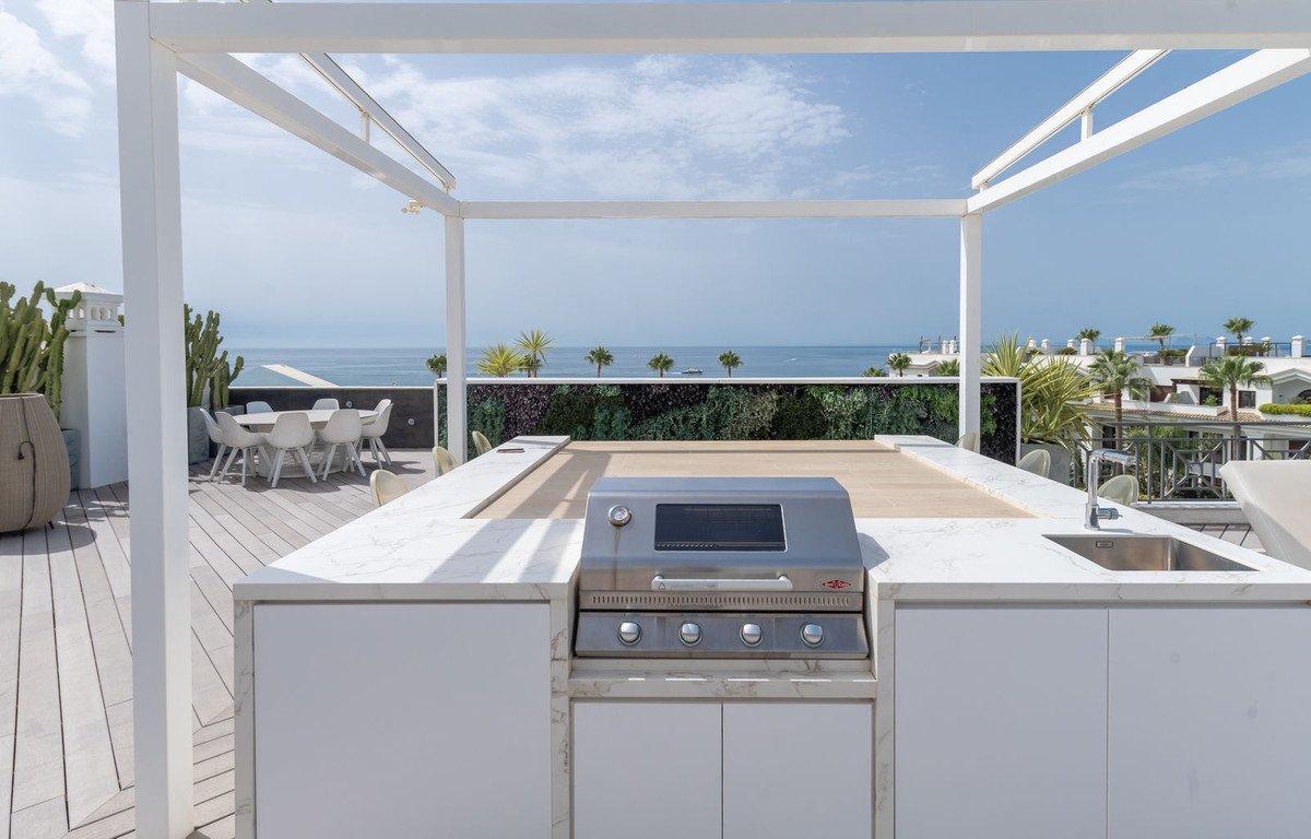 Modern rooftop kitchen with grill, sink, and ocean view, featuring white cabinetry and outdoor dining area.