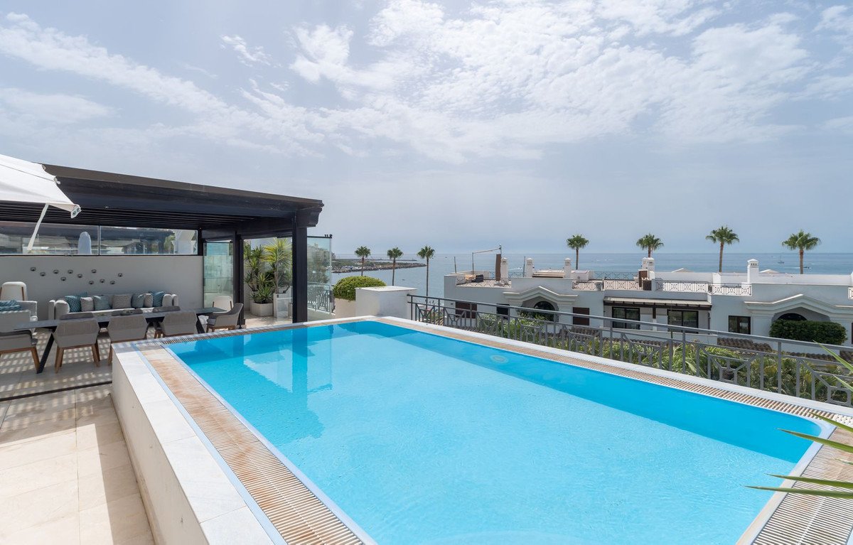 Rooftop pool with ocean view, lounge area, and palm trees under a cloudy sky.