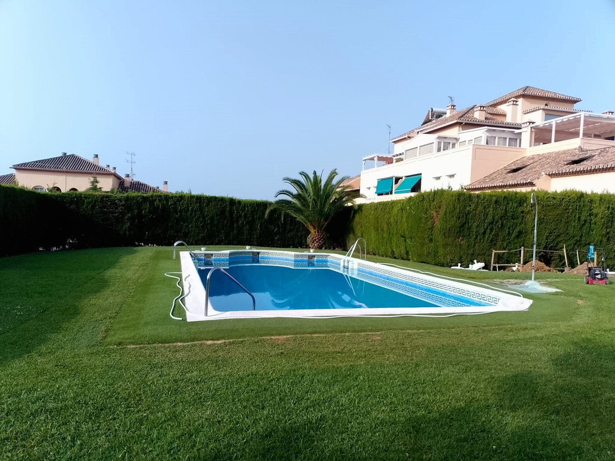 A beautiful swimming pool with blue water is surrounded by a well-maintained green lawn and tall hedges, with villas in the background.