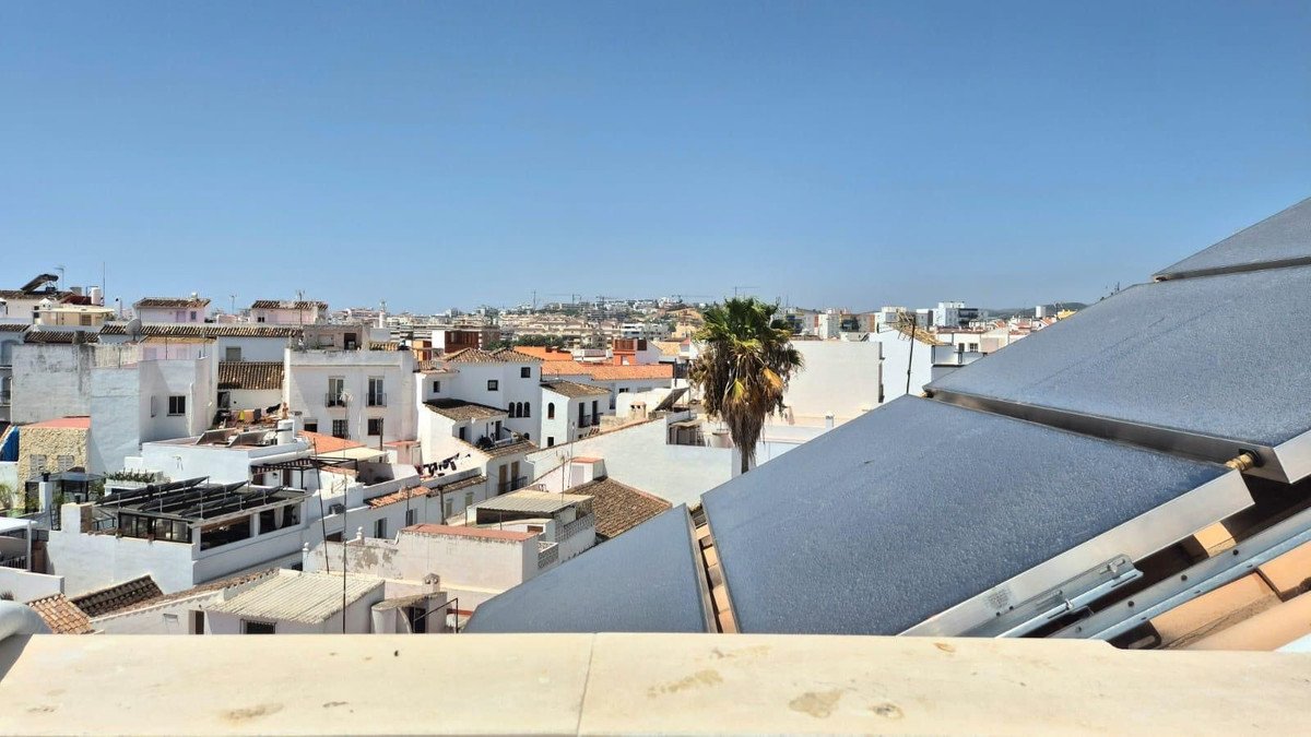 View over a city with white buildings and a palm tree, under a clear blue sky. Rooftop solar panels are visible.