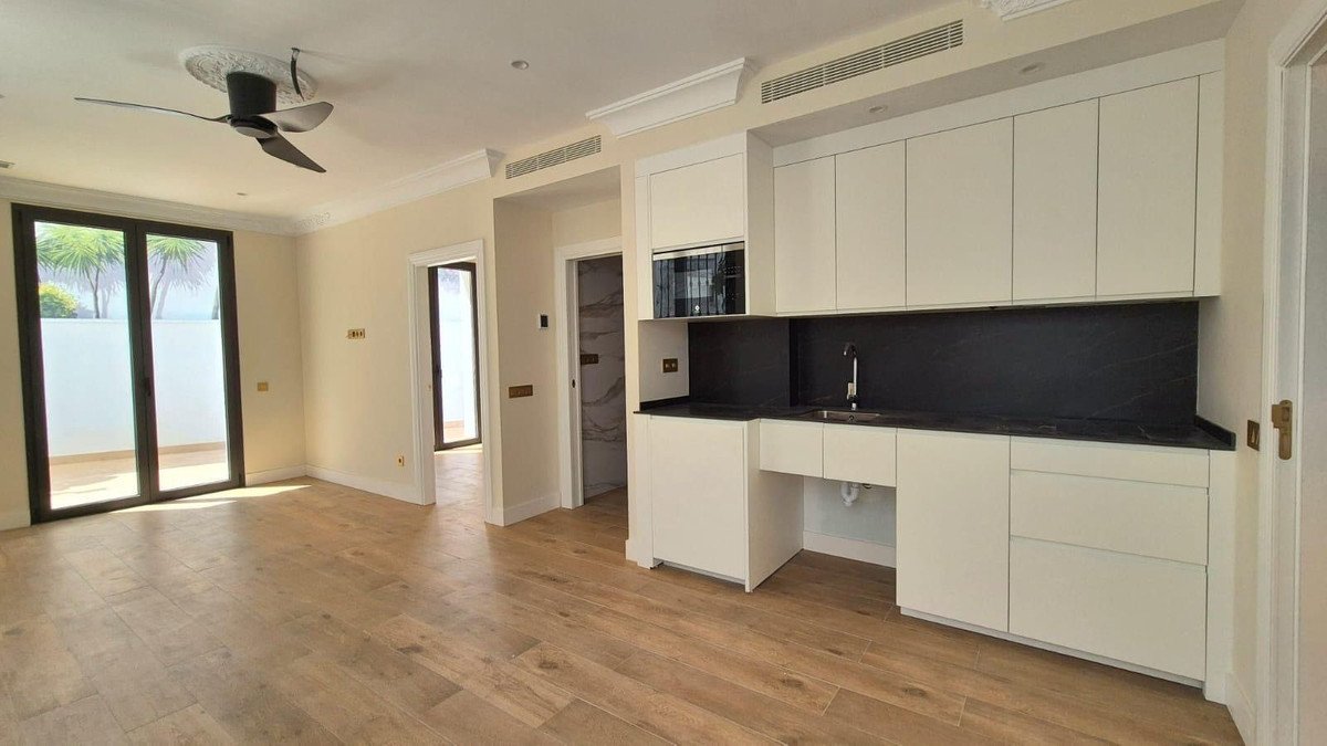 Modern white kitchenette with black countertop and microwave, connected to a spacious living area with terrace access and palm trees outside.