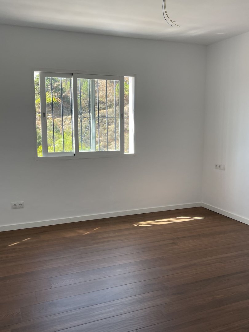 Empty room with wooden flooring, white walls, and a window offering a view of greenery outside.