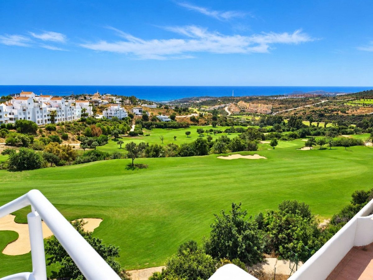 View of a golf course with sand traps, trees, houses, and the ocean in the distance under a blue sky.