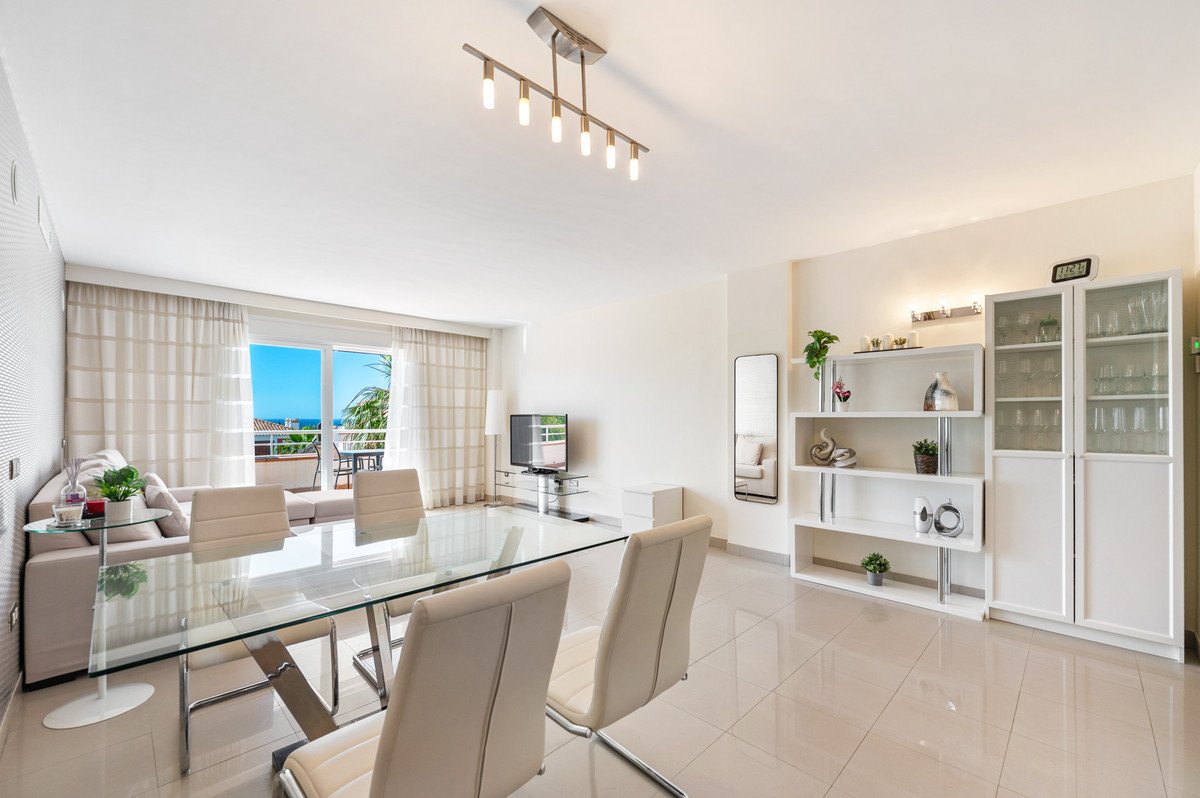Modern living room with glass dining table, white furniture, and ocean view through large windows.