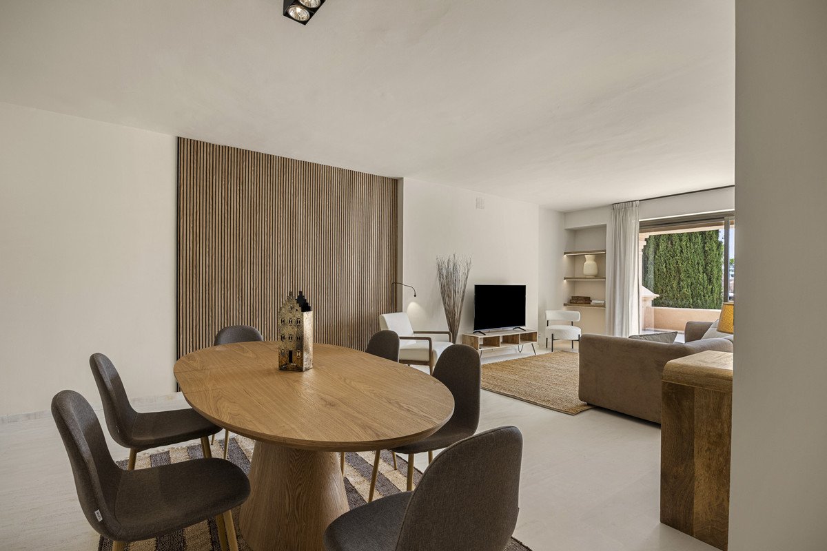 Modern living room featuring a wooden dining table with chairs, a sofa, and a TV in a bright space with large windows