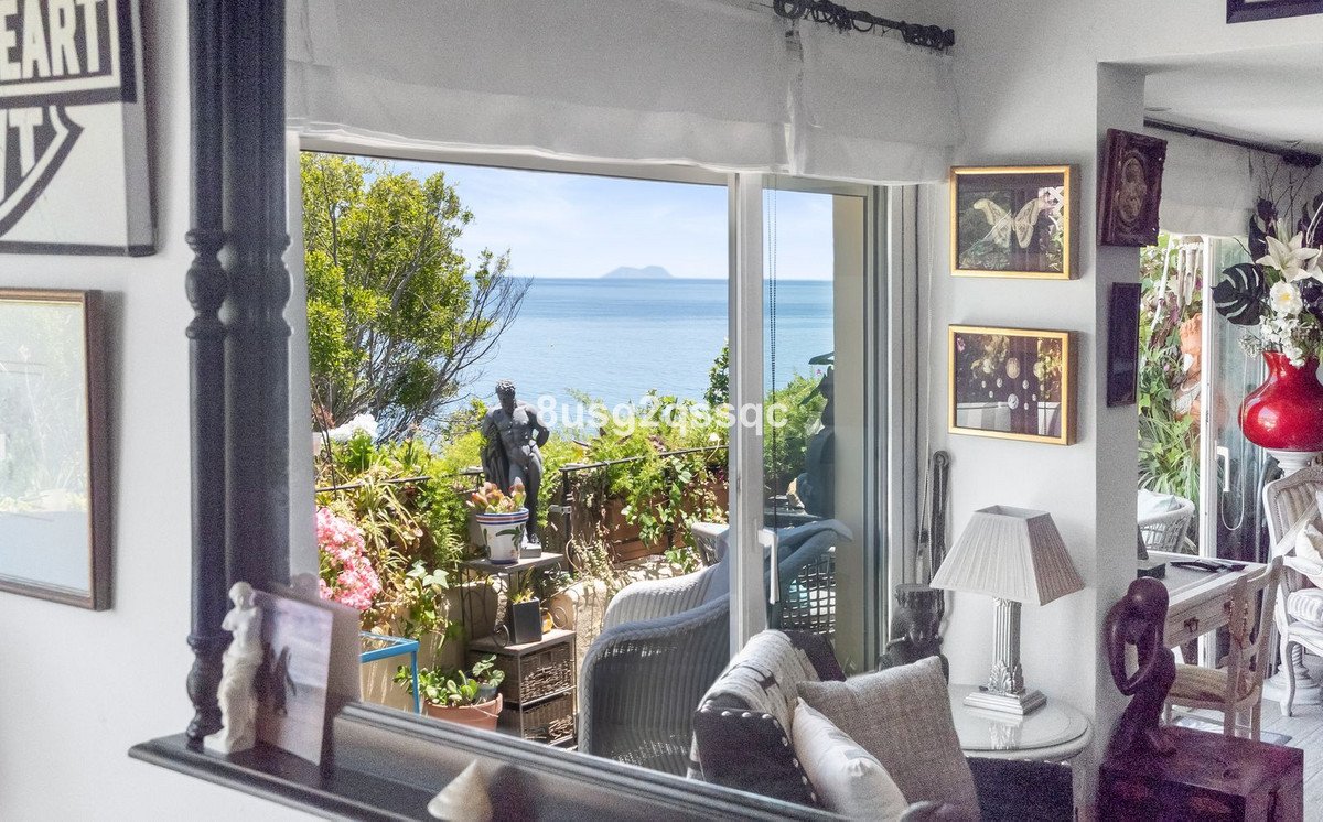 Living room overlooking the sea with a balcony featuring lush greenery and statues, offering a picturesque ocean view.