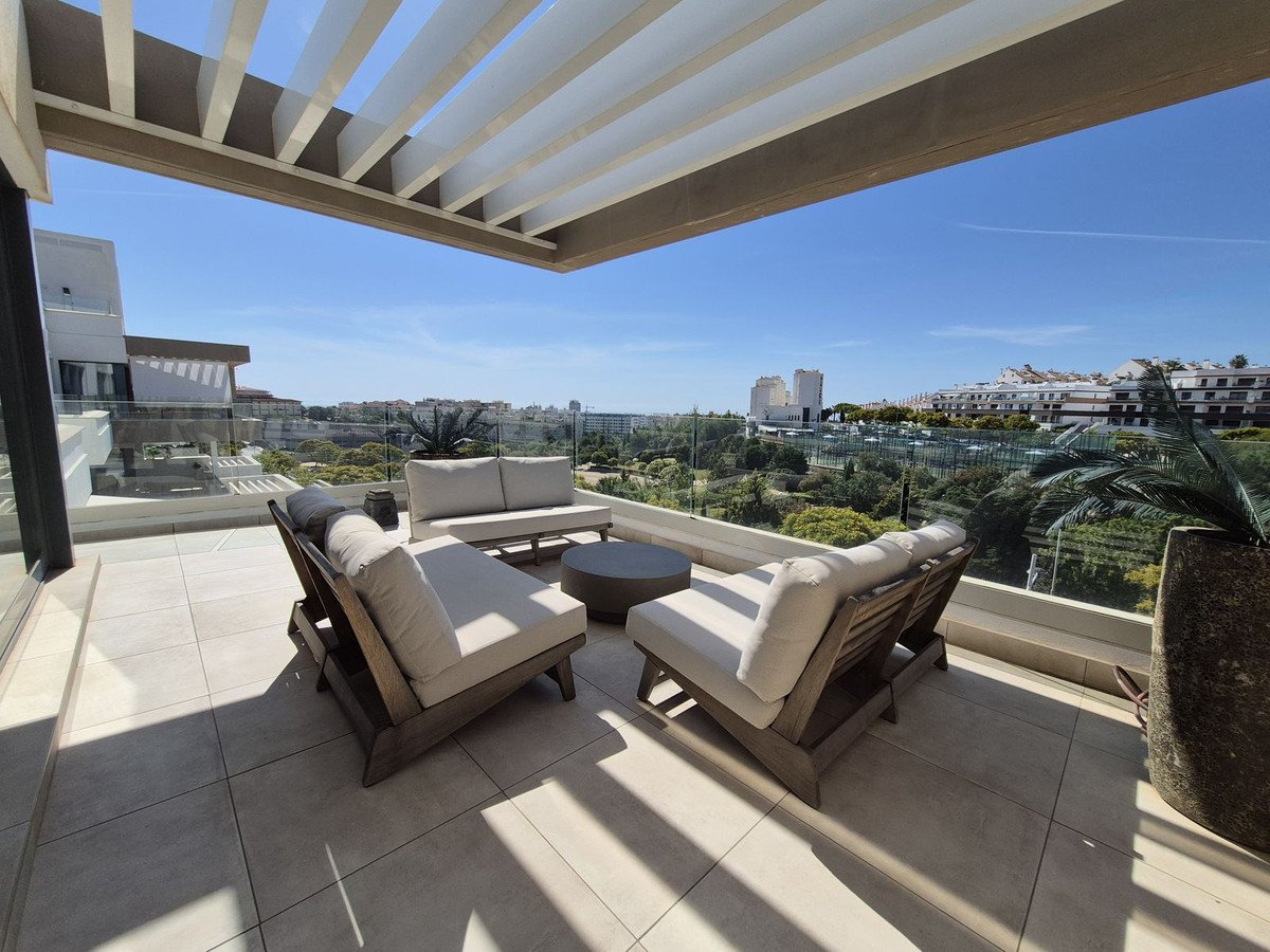 Spacious terrace with modern lounge furniture and a panoramic view of lush greenery and distant buildings under a clear blue sky.