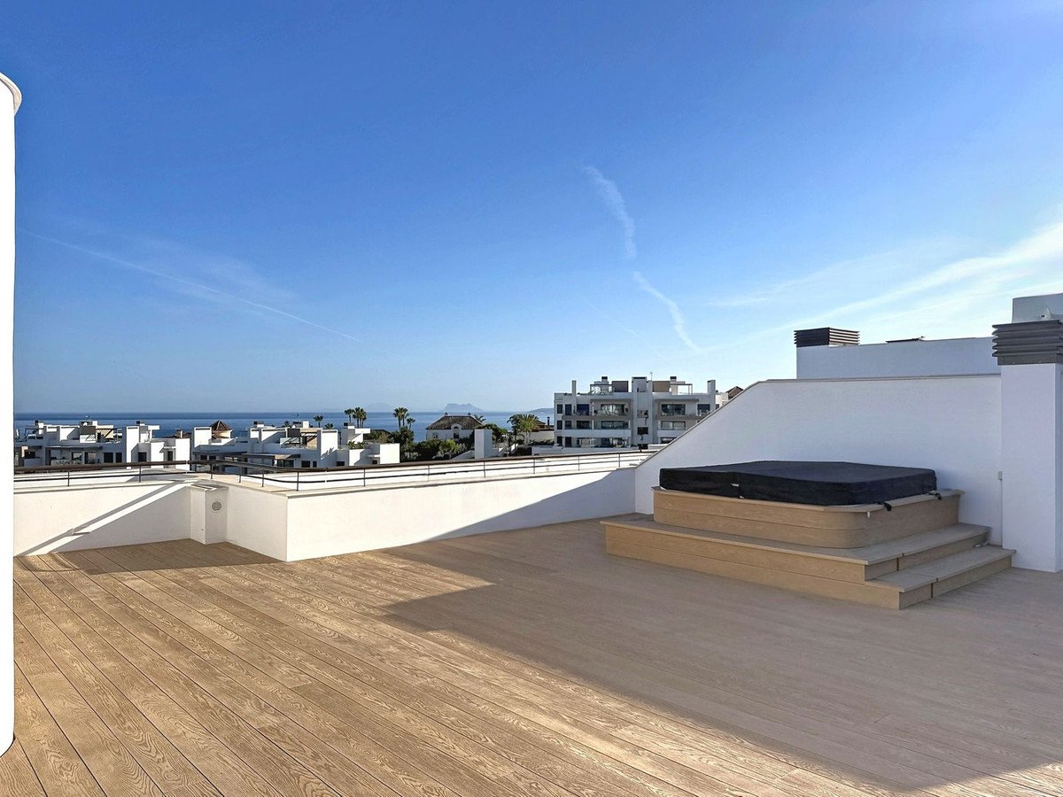 Rooftop terrace with wooden deck, hot tub, and ocean view under clear blue sky.