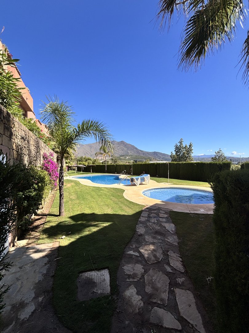 Two sparkling blue swimming pools set in a lush green garden with a mountain range visible in the distance.