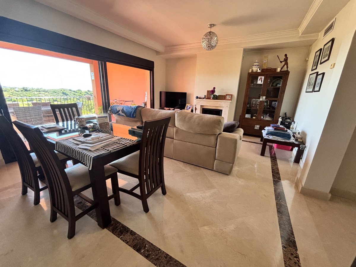 Spacious living and dining area with a glass-top table, comfortable sofa, fireplace, and a view of the balcony and greenery.