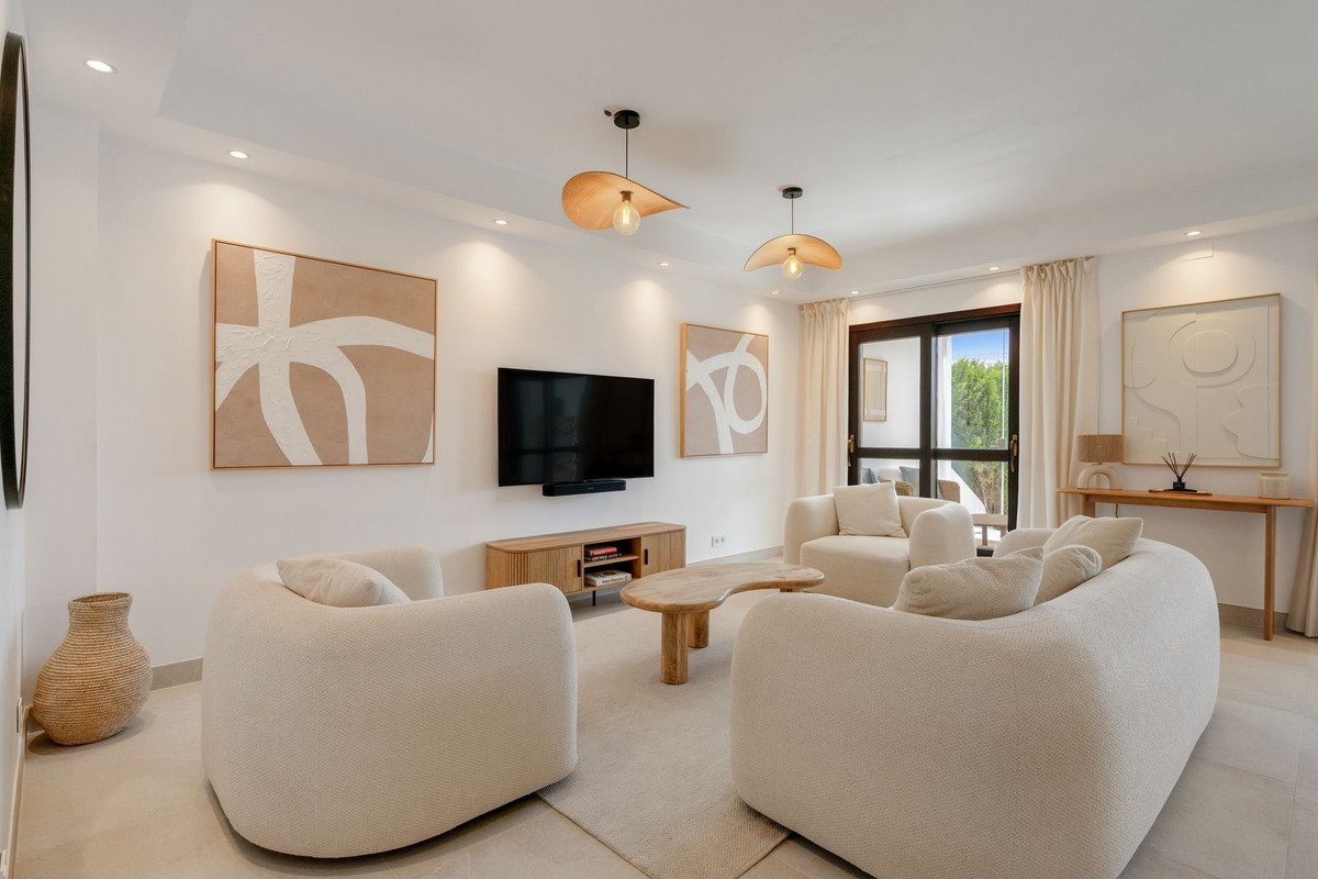 Modern living room with white sofas, wooden furniture, abstract art, and large windows overlooking greenery.