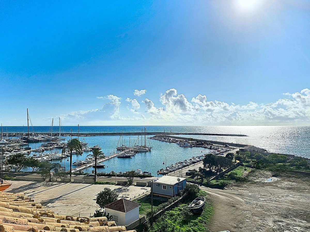 A scenic view of a marina filled with boats, overlooking the calm sea under a bright blue sky with scattered clouds.