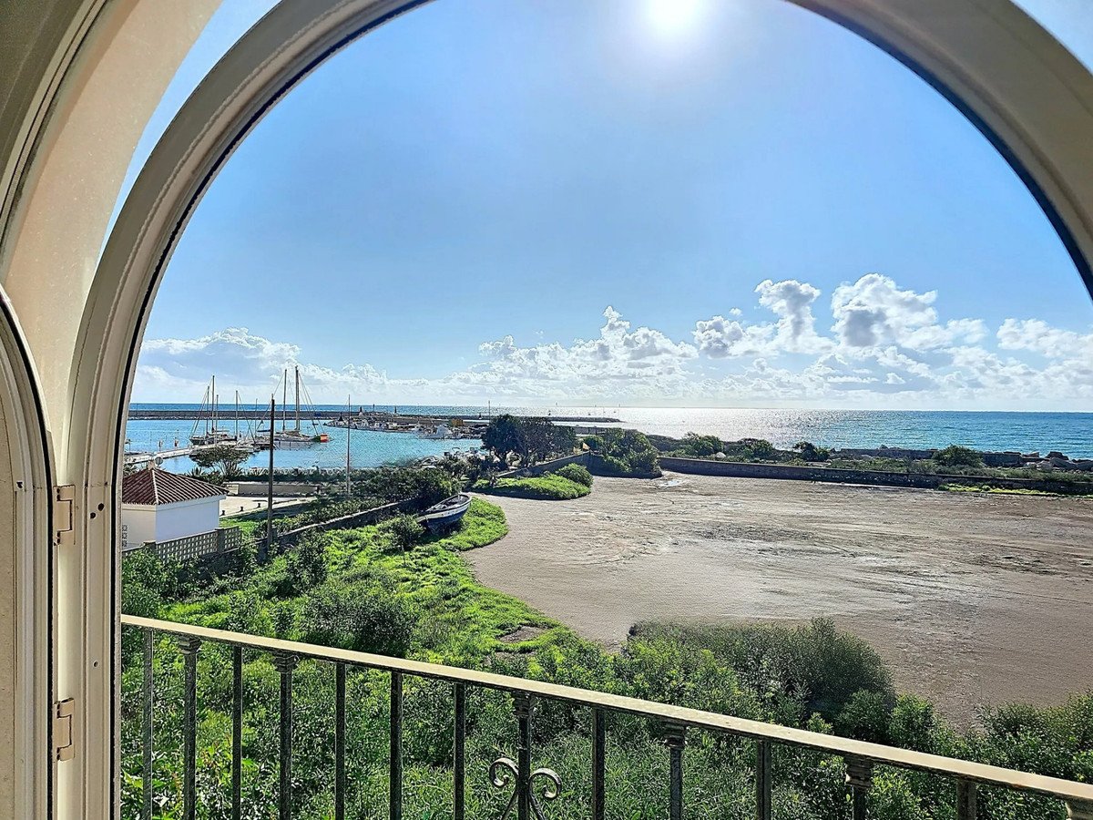 View from a balcony through an arched window of a yacht harbor with boats and the blue sea under a sunny sky.