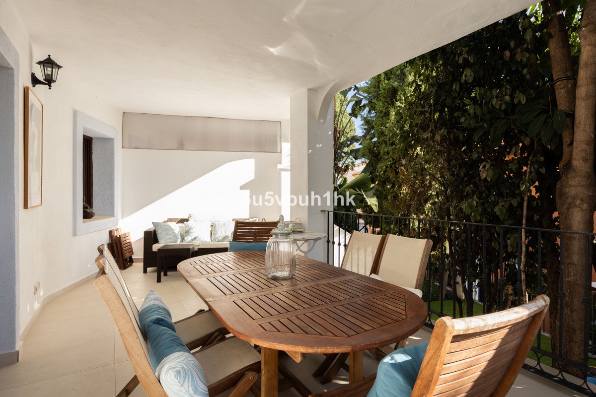 Covered terrace with wooden dining table, chairs, and outdoor sofa, surrounded by lush greenery.