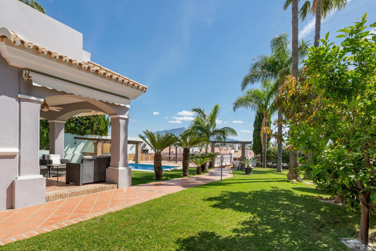 Covered patio with outdoor seating next to a swimming pool surrounded by palm trees and lush gardens, with mountains in the background.