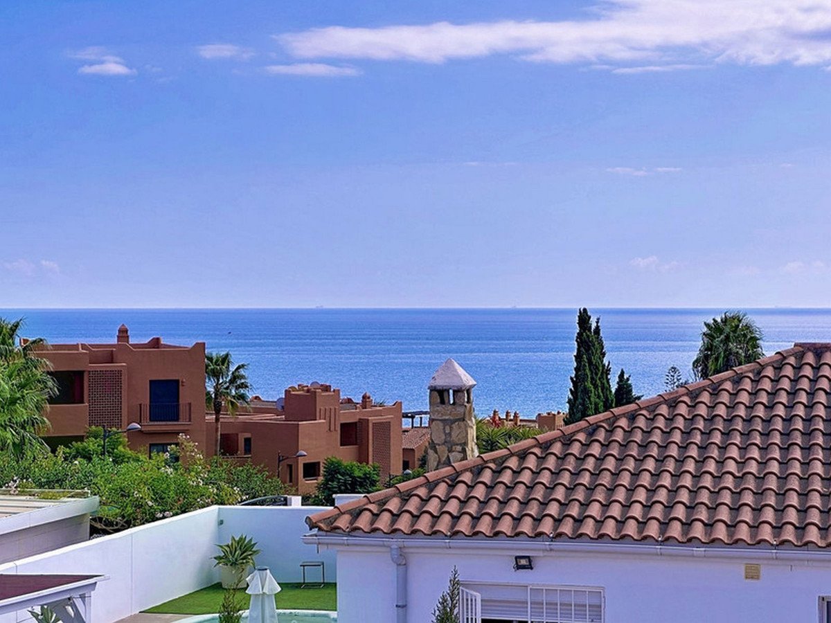 A scenic view overlooking terracotta tiled roofs towards the blue sea under a clear sky with scattered clouds.