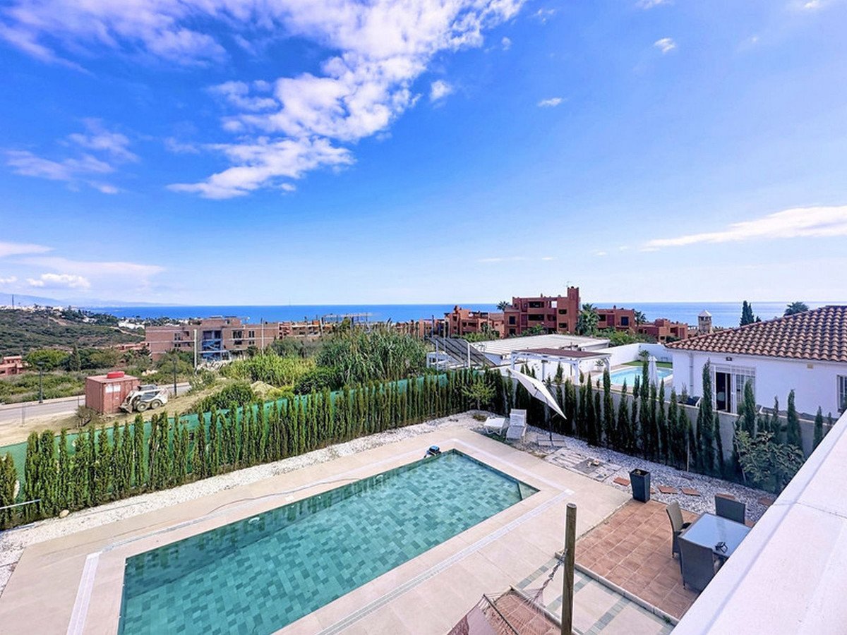 A villa with a swimming pool and outdoor seating area, overlooking the sea and surrounding hills under a blue sky.