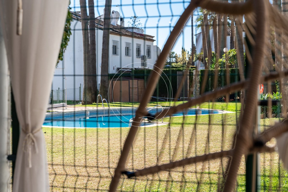 A view of a swimming pool and garden through a fence, with palm trees and a building in the background.