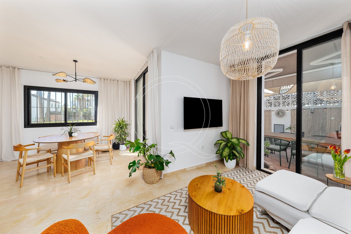 Bright living and dining area with large windows, modern furniture, plants, and a view of an outdoor patio.
