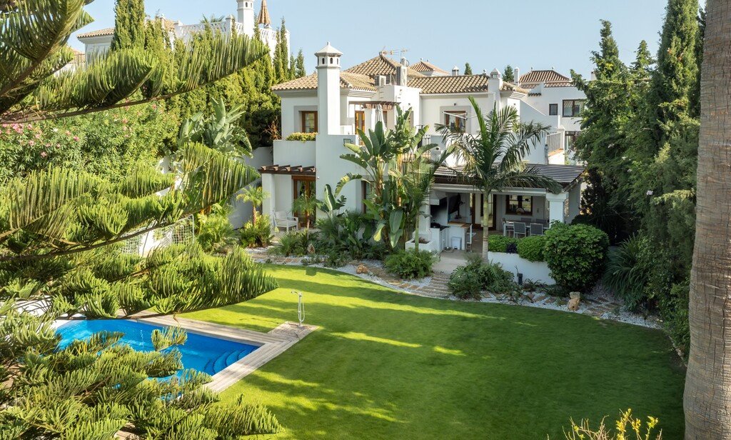 A white villa with a swimming pool, lush green garden, and tropical plants under a clear blue sky.