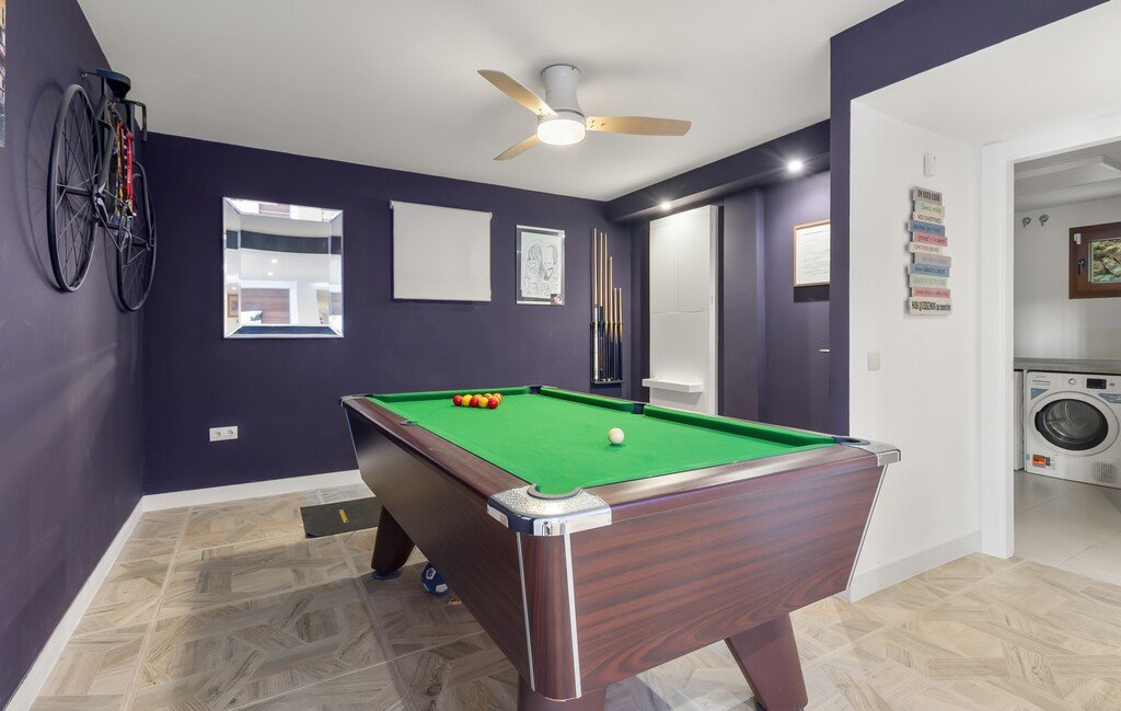 A pool room with a billiard table, bicycle mounted on the wall, and a ceiling fan. A laundry area is visible through a doorway.