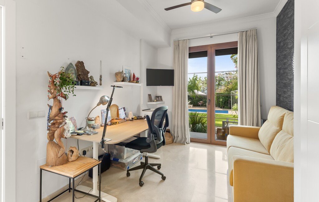 Home office with a desk, chair, and a sofa, featuring a large window overlooking a backyard with a swimming pool.