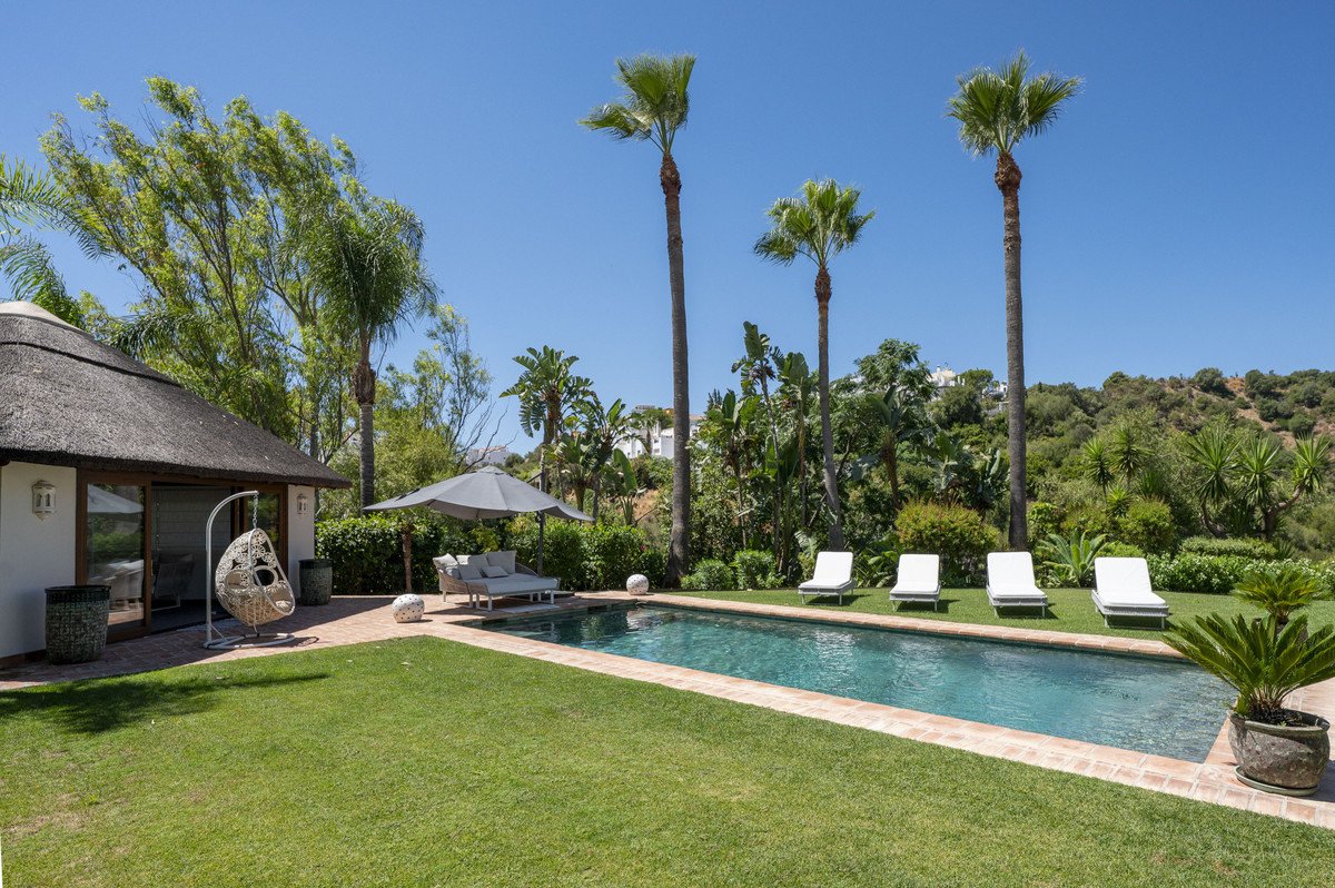 Luxurious outdoor area with a swimming pool, lounge chairs, sofa, and a thatched-roof structure on a sunny day.