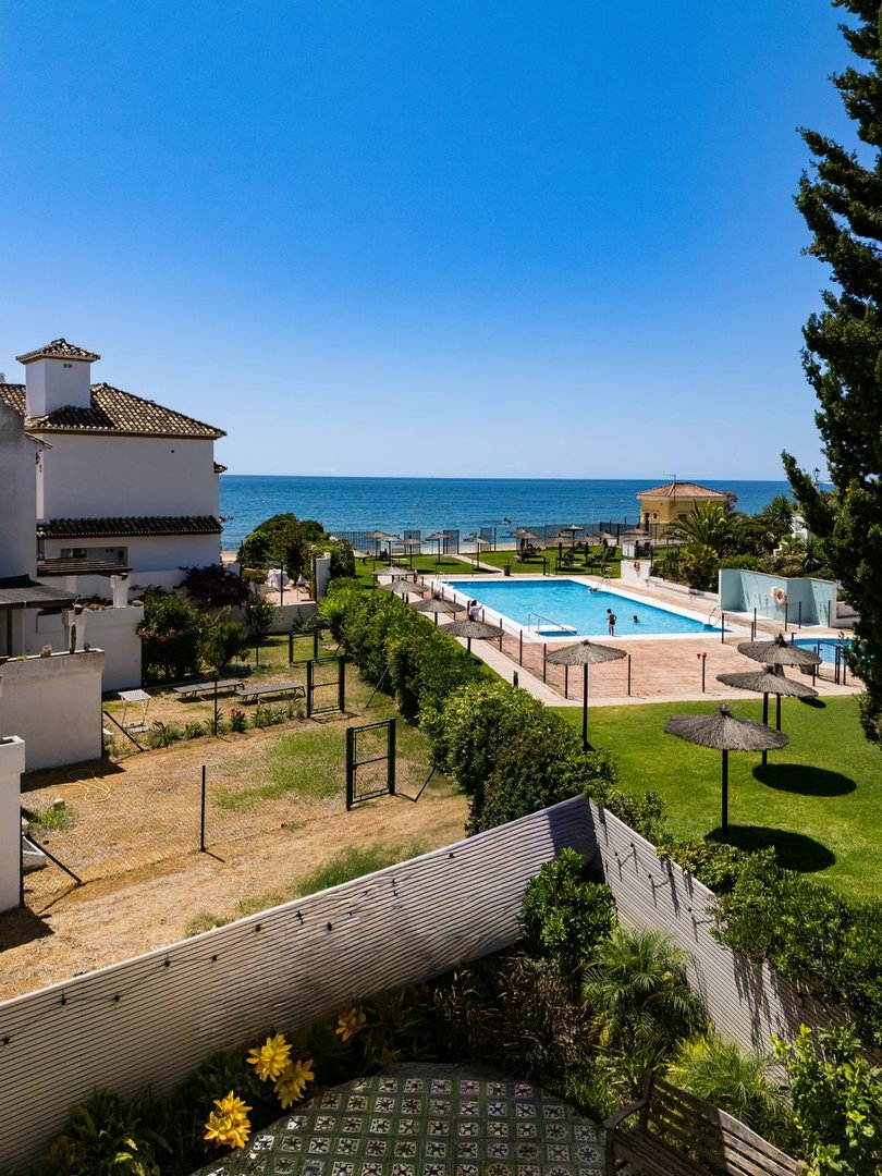 Sea view from a balcony overlooking a pool, garden, and ocean under a clear blue sky.