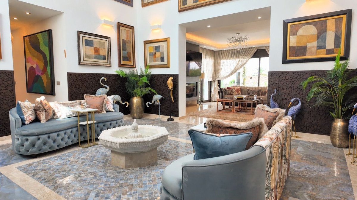 Elegant living room with plush seating, decorative sculptures, a central fountain, and artistic wall decor, leading to another seating area.