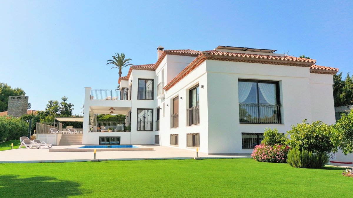 A large white villa with a swimming pool, manicured lawn, and outdoor seating area on a sunny day.