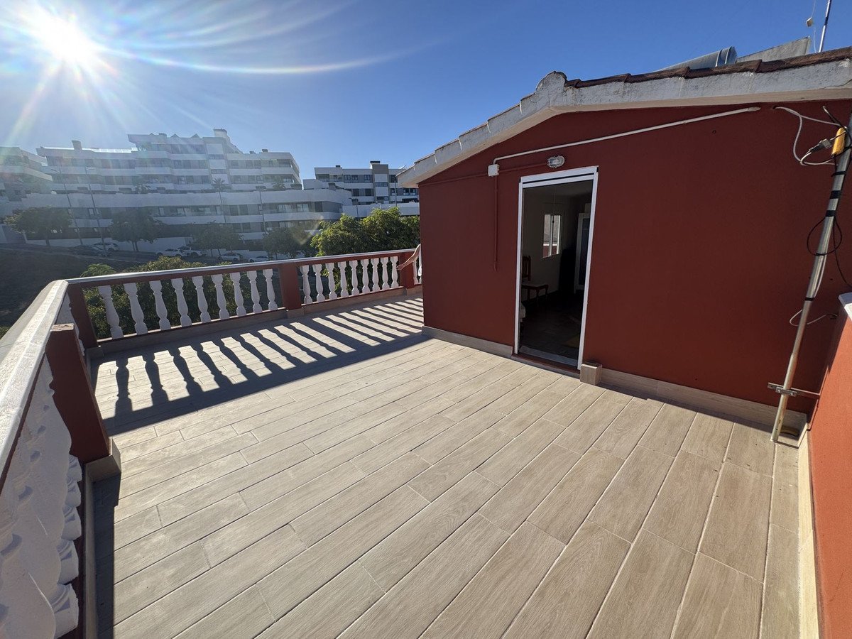 Spacious terrace with wooden flooring and a railing, offering views of buildings under a sunny sky.