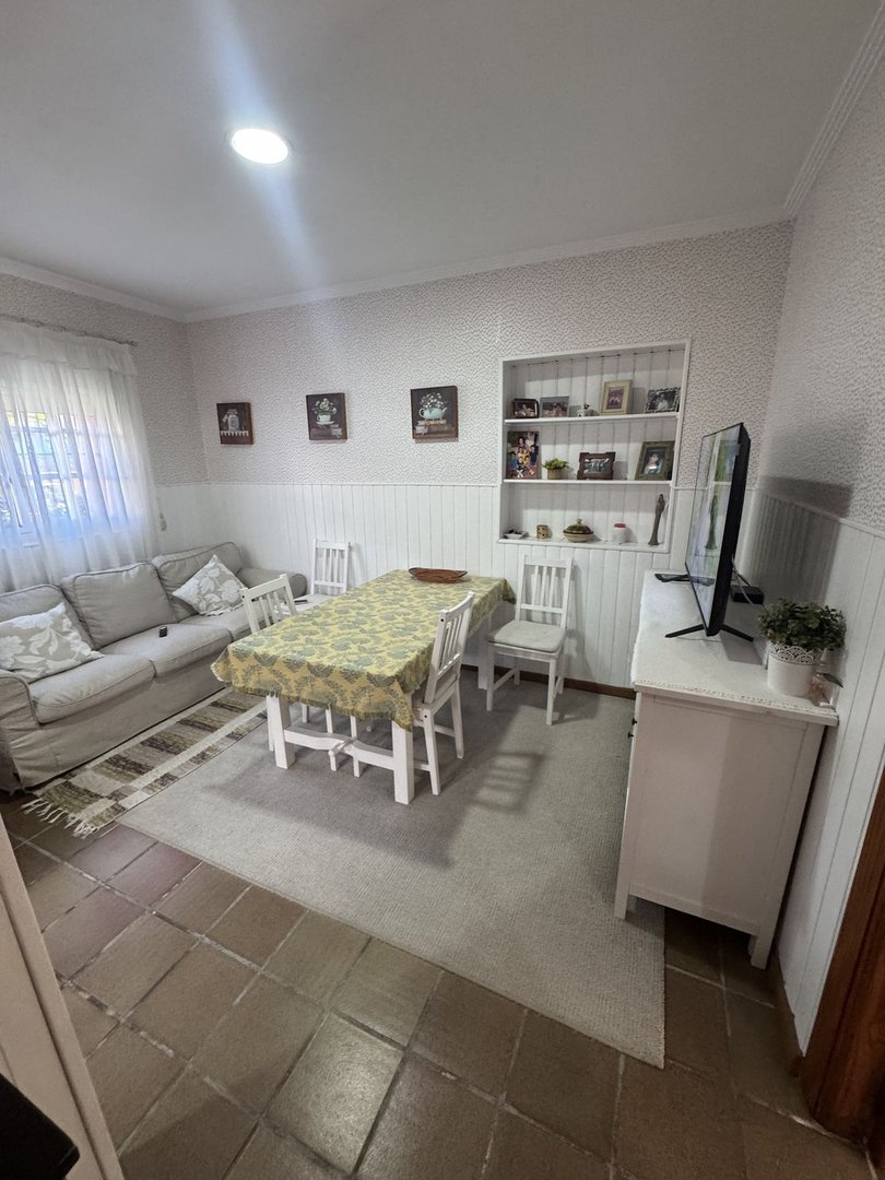 A dining and living area with a sofa, dining table, chairs, a TV, and shelves. Features a patterned tablecloth.