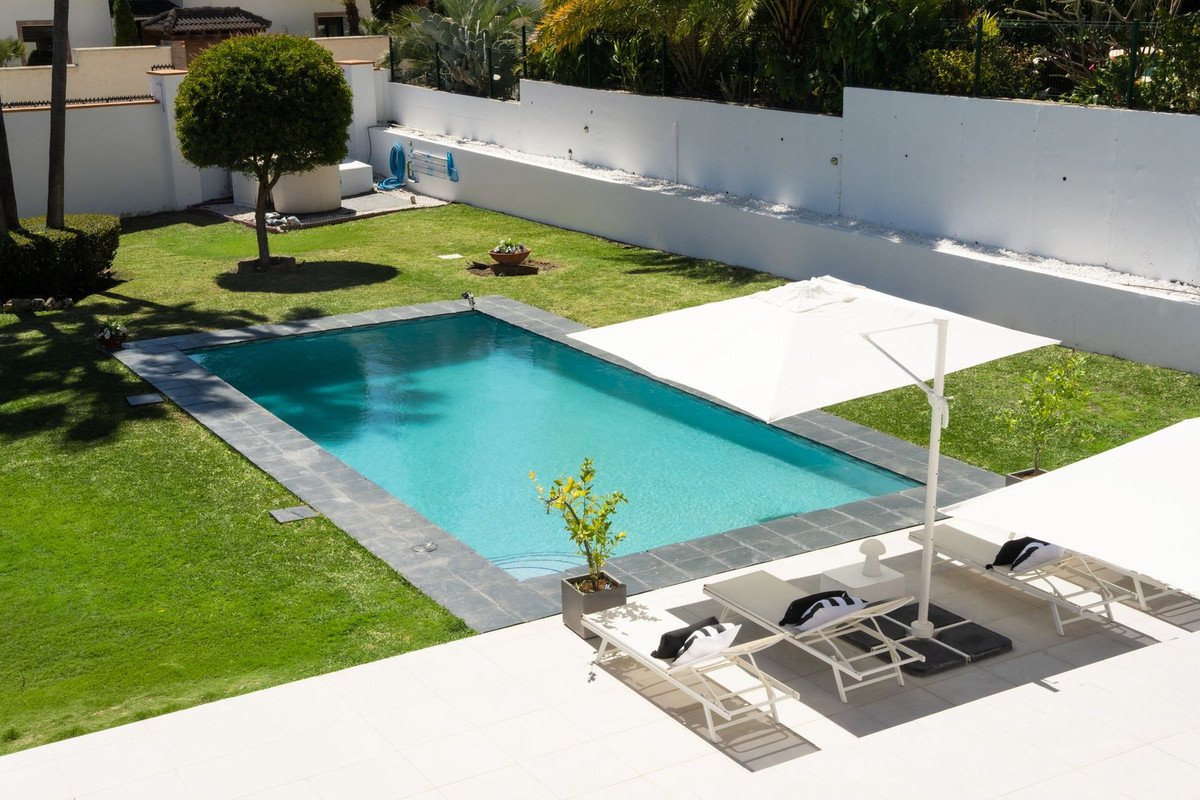 A rectangular swimming pool surrounded by a lush green lawn, lounge chairs, and white umbrellas, with a garden and trees in the background.