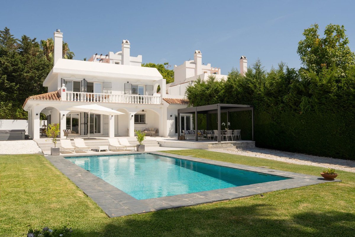 A white villa with a swimming pool, sun loungers, and a covered outdoor dining area, surrounded by lush green trees.