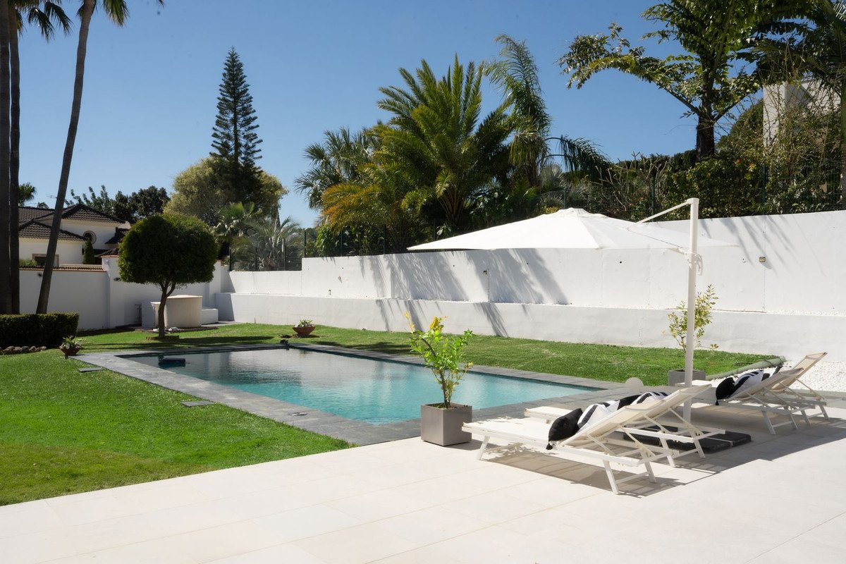 A spacious outdoor area with a rectangular swimming pool, surrounded by green grass, white lounge chairs, and lush palm trees under a clear blue sky.