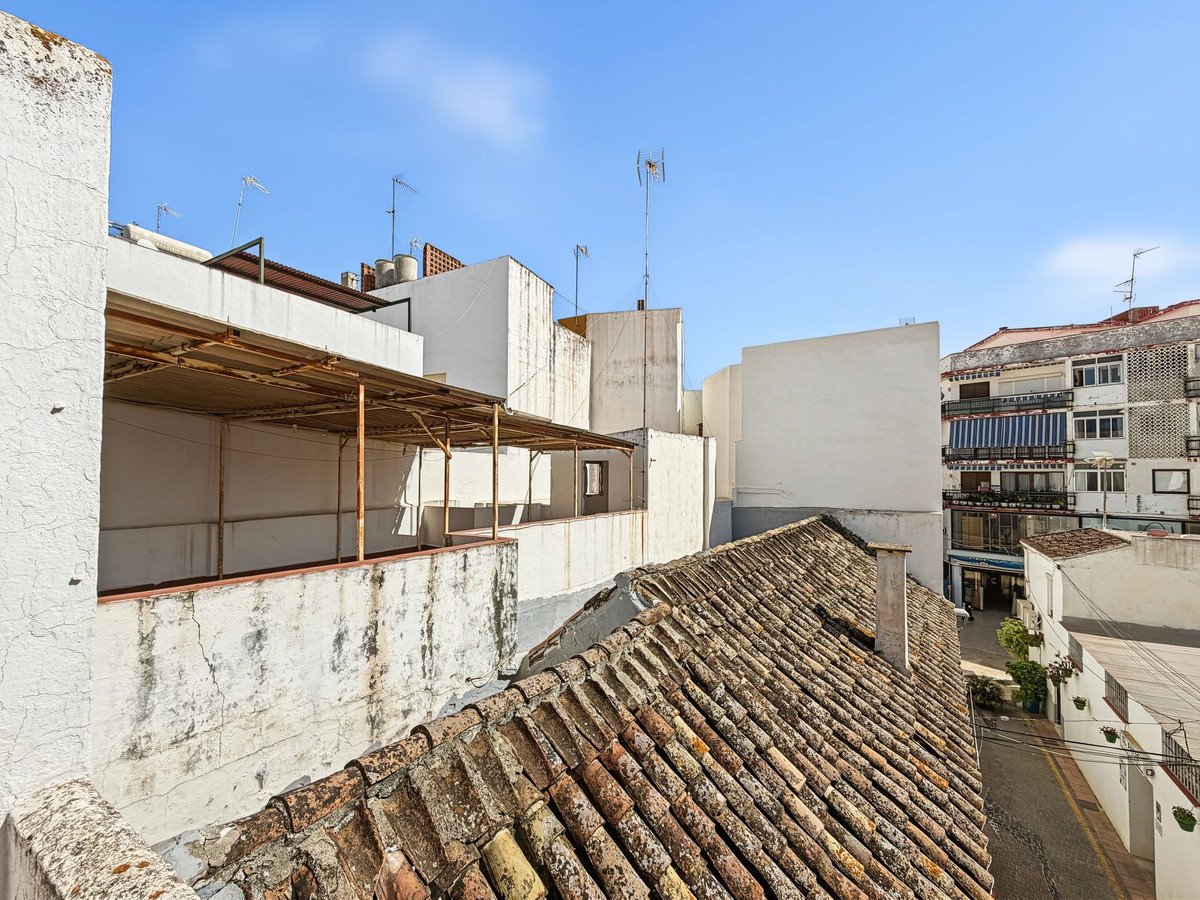 View from a rooftop overlooking a street, showing neighboring buildings with balconies and tiled roofs under a clear blue sky.