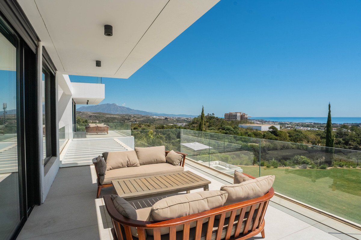 Modern balcony with wooden furniture, glass railings, and panoramic views of the sea, mountains, and green landscape.