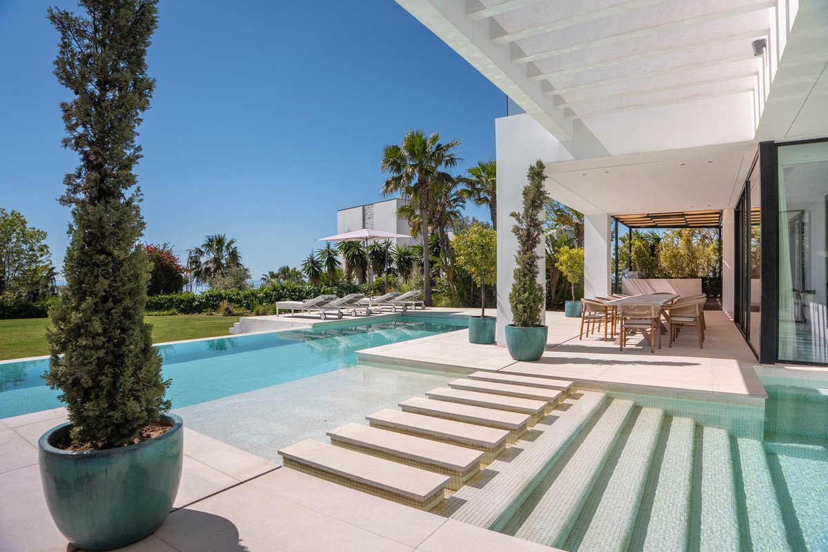 Modern villa with infinity pool, sun loungers, and ocean view under clear blue sky.