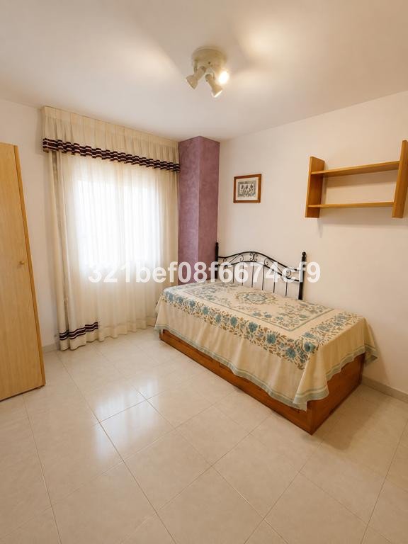 A bedroom with a single bed, floral blanket, sheer curtains, and wooden shelves on the wall.