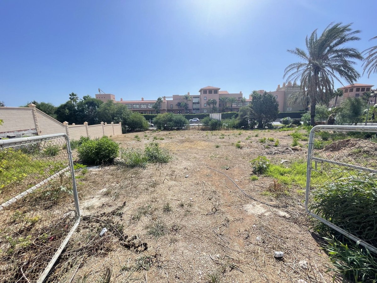 Vacant desert lot with a fence and buildings in the background under a clear blue sky.