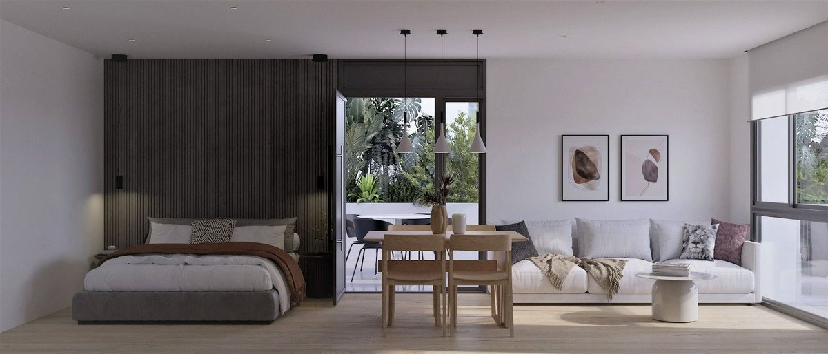 A modern studio apartment with a bed, living area, dining table, and balcony access overlooking lush greenery.