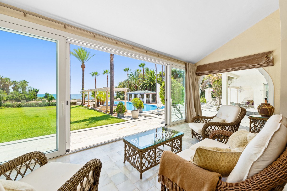 Sunroom with wicker furniture overlooking a lush garden, pool, and the sea with palm trees.