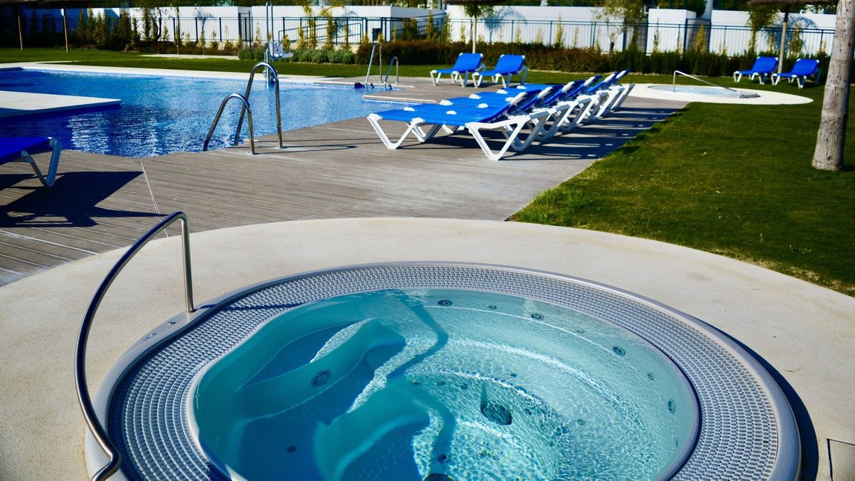 An outdoor area with a hot tub in the foreground and a swimming pool with lounge chairs in the background.