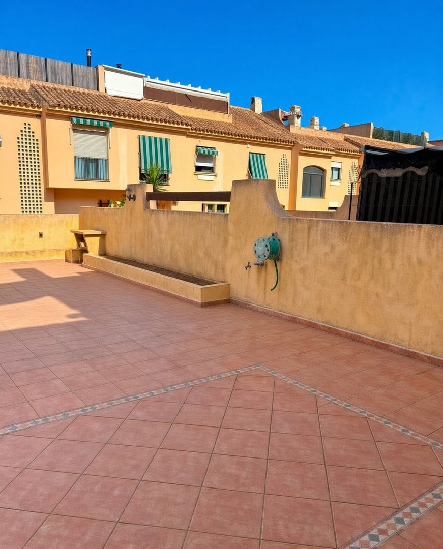 A sunny terrace with terracotta tiles and a long planter against an orange building under a clear blue sky.