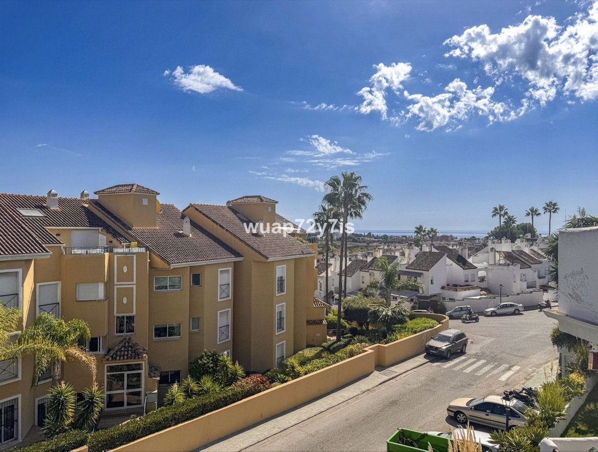 View from a balcony of apartments with palm trees and a distant view of the sea under a blue sky with clouds.