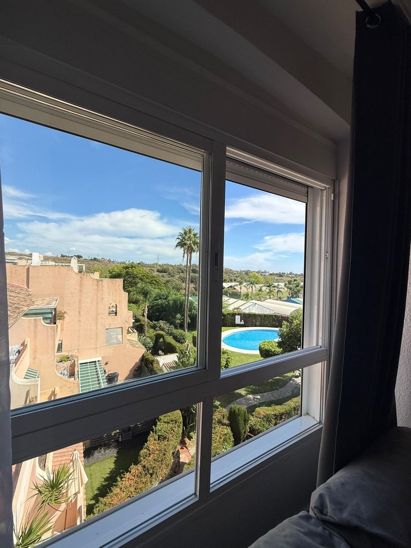 View from a window overlooking a swimming pool, lush greenery, and apartment buildings on a sunny day.