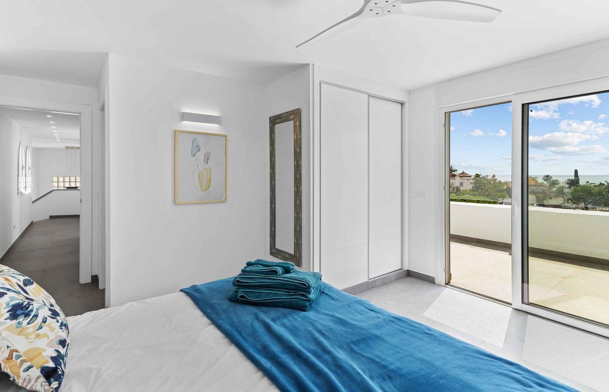 A bedroom with a balcony door offering a sea view, a bed with blue throws, and a ceiling fan.