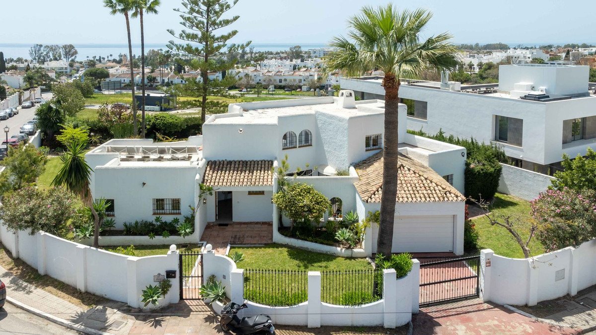 A white villa with a tile roof, surrounded by palm trees, green lawn, and a white wall, with a distant view of the sea.