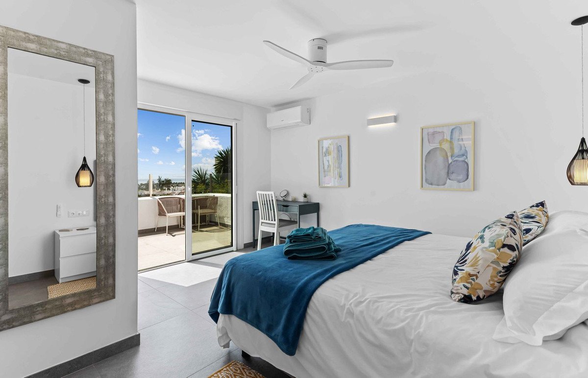 Bedroom with a large bed, desk, and balcony overlooking the sea and palm trees.