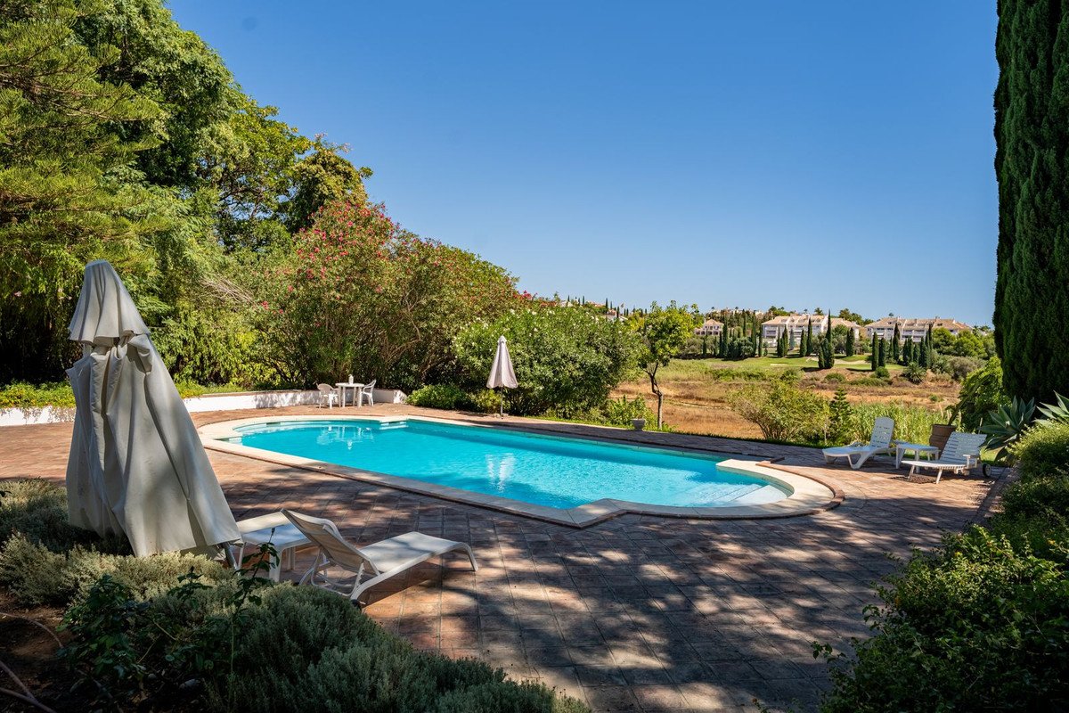 Outdoor swimming pool with lounge chairs and a distant view of buildings and greenery under a clear blue sky.